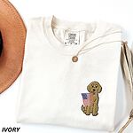 Ivory T-shirt folded with a necklace, a sitting golden doodle with a stars-and-stripes flag stitched on the left chest