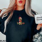 Woman holding a mug in a Black crewneck sweatshirt embroidered with a golden doodle in a Santa hat and scarf with gifts