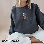 Woman sitting in a Dark Heather crewneck sweatshirt embroidered with a cream doodle in a Santa hat and red scarf with gifts