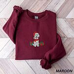 Maroon crewneck sweatshirt laid flat on a wood floor, embroidered with a cream doodle in a Santa hat and scarf between two gifts