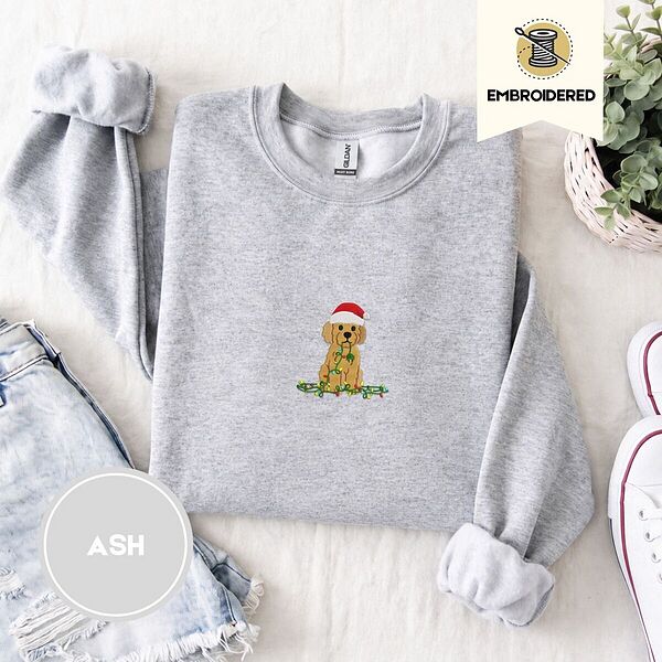 Ash crewneck sweatshirt folded with denim shorts, white sneakers and a potted plant, embroidered with a goldendoodle puppy in a Santa hat chewing on a tangled string of Christmas lights