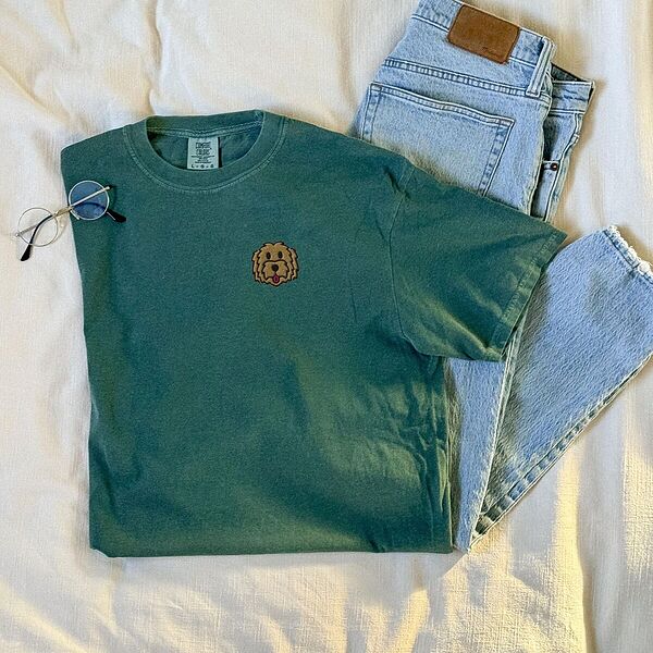 Blue Spruce T-shirt laid flat on a bed beside jeans and glasses, embroidered with a golden doodle face on the left chest