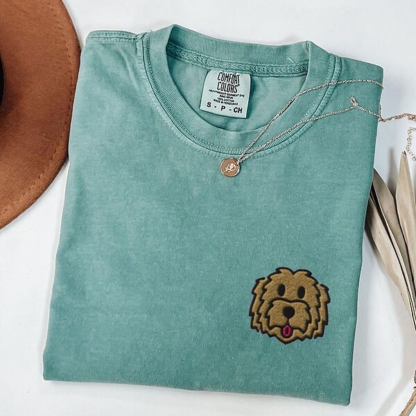 Light Green T-shirt laid flat with a hat and a necklace, embroidered with a shaggy golden doodle face on the left chest