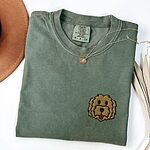 Moss T-shirt laid flat with a hat and a necklace, embroidered with a shaggy golden doodle face on the left chest