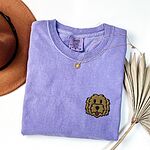 Violet T-shirt laid flat with a hat and dried palm leaves, embroidered with a shaggy golden doodle face on the left chest