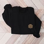 Black crewneck sweatshirt laid flat on wood, embroidered with a small shaggy golden doodle face on the left chest