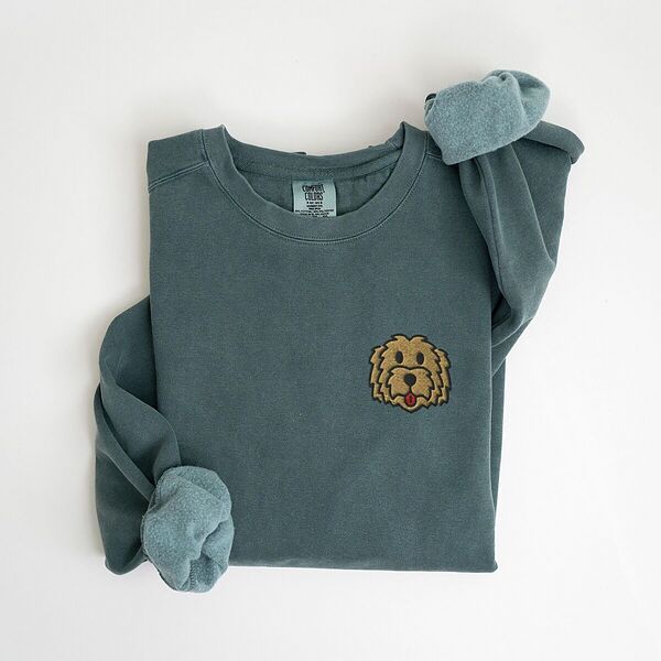 Blue Spruce crewneck sweatshirt laid flat with a rolled cuff, embroidered with a small shaggy golden doodle face on the left chest