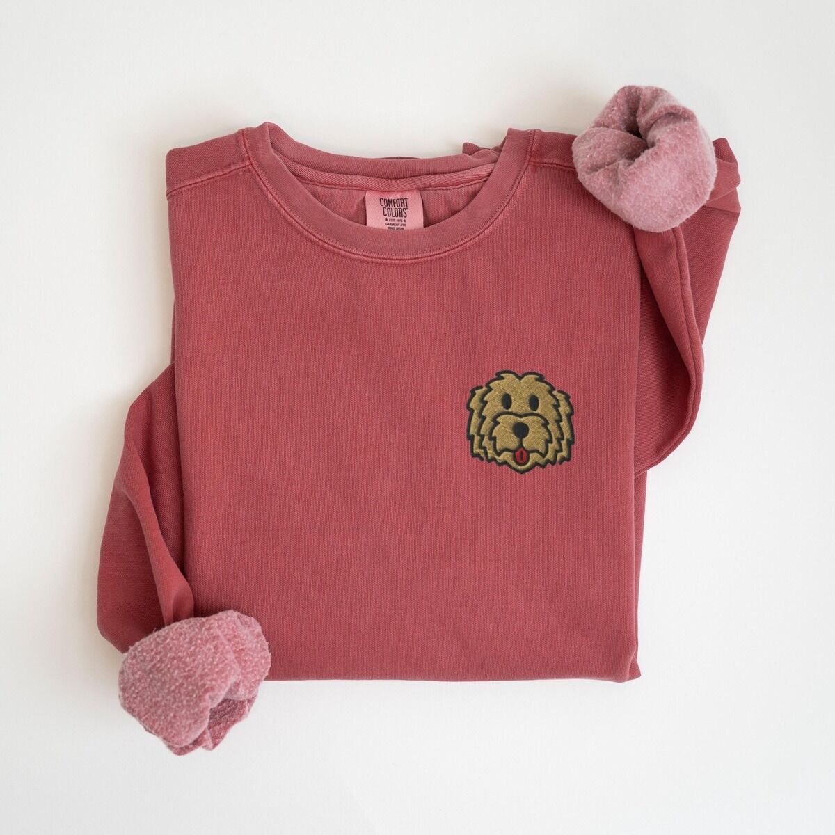 Goldendoodle Face Sweatshirt – Crimson Crimson crewneck sweatshirt laid flat with a knotted sleeve, embroidered with a small shaggy golden doodle face on the left chest