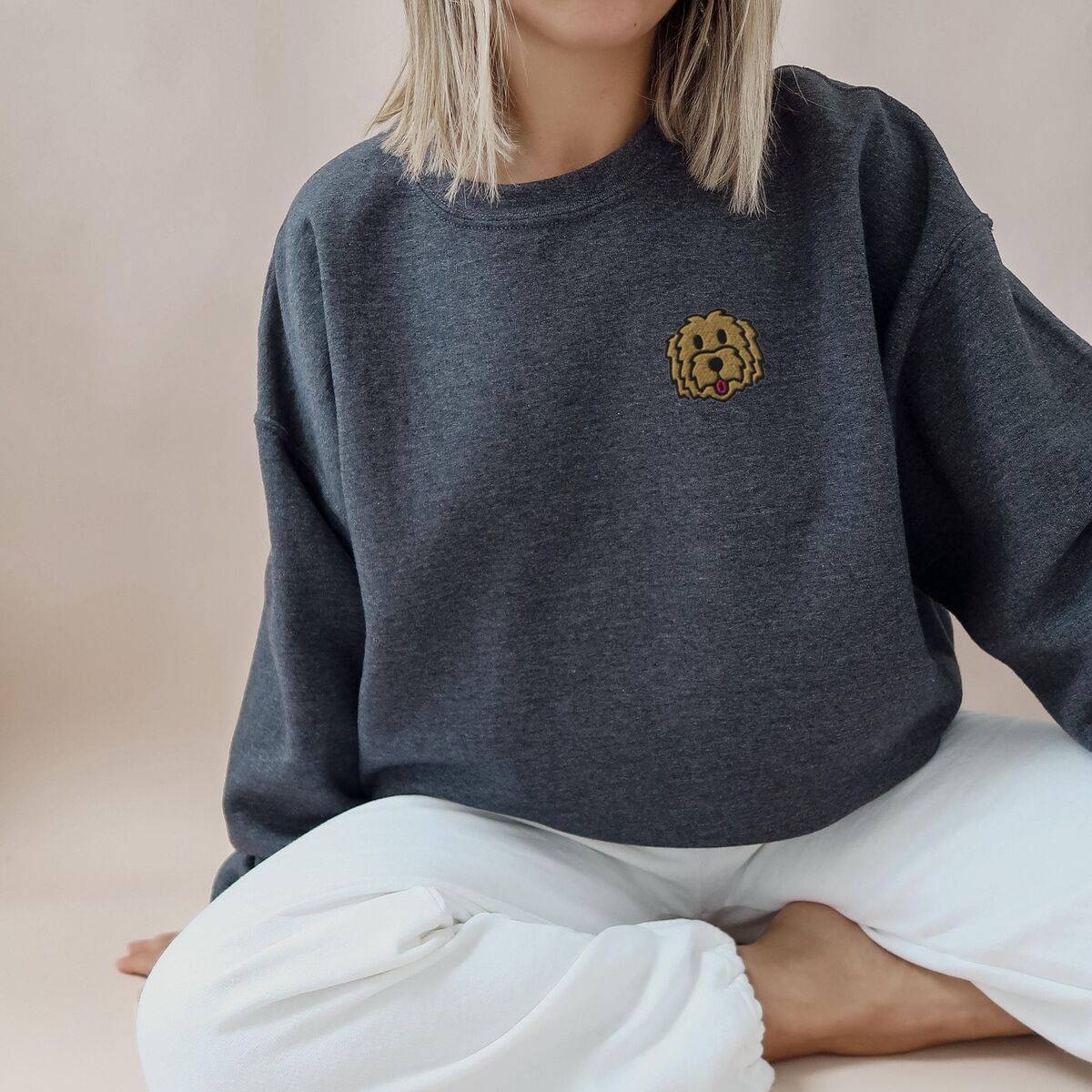 Goldendoodle Face Sweatshirt - Dark Heather Woman sitting in a Dark Heather crewneck sweatshirt embroidered with a small golden doodle face on the left chest
