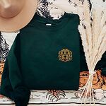 Forest Green crewneck sweatshirt laid flat with a hat and pampas grass, embroidered with a small shaggy golden doodle face on the left chest
