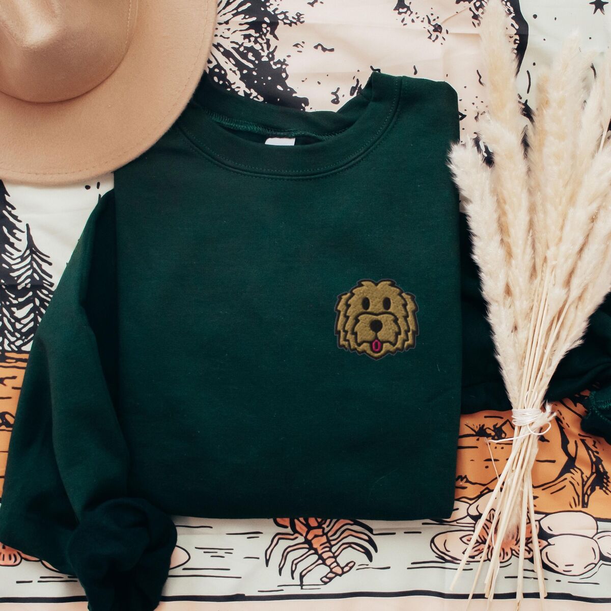 Goldendoodle Face Sweatshirt - Forest Green Forest Green crewneck sweatshirt laid flat with a hat and pampas grass, embroidered with a small shaggy golden doodle face on the left chest