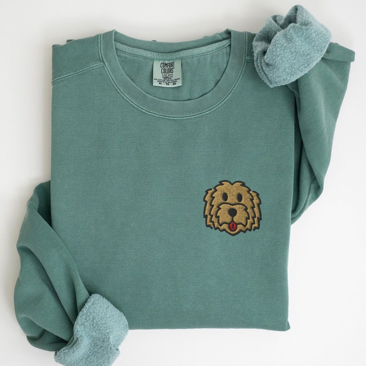 Goldendoodle Face Sweatshirt – Light Green Light Green crewneck sweatshirt laid flat with a rolled cuff, embroidered with a small shaggy golden doodle face on the left chest