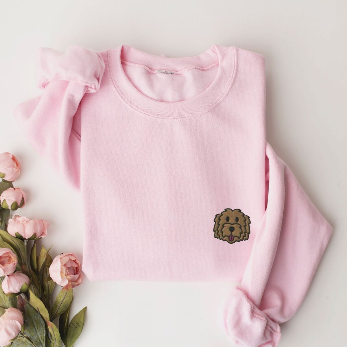 Goldendoodle Face Sweatshirt - Light Pink Light Pink crewneck sweatshirt laid flat beside pink roses, embroidered with a small shaggy golden doodle face on the left chest