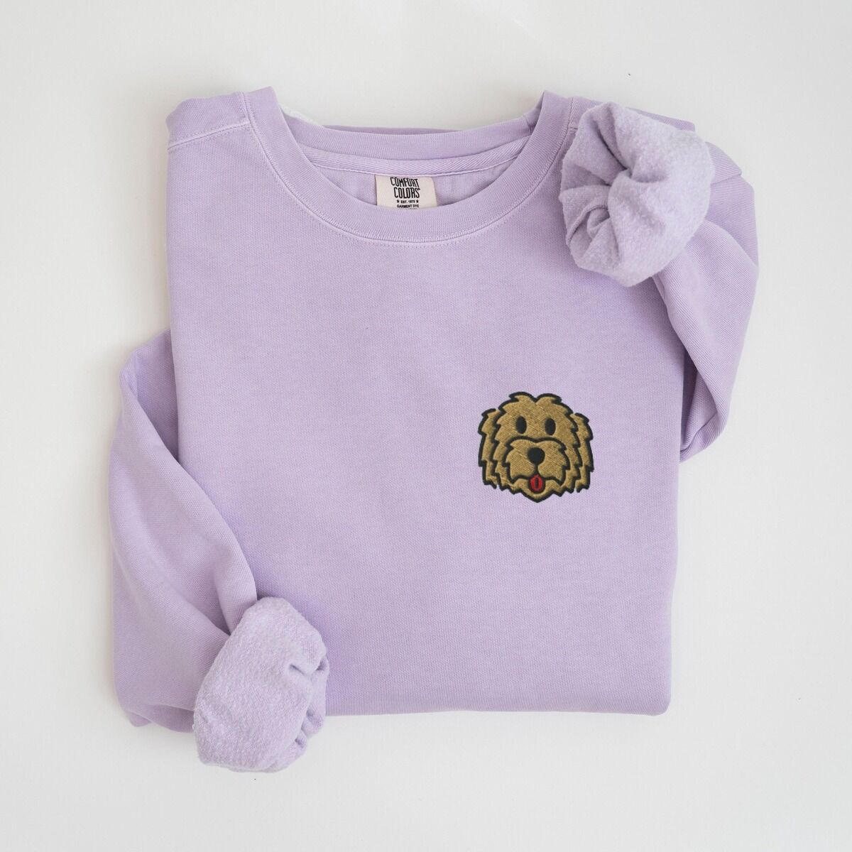 Goldendoodle Face Sweatshirt – Orchid Orchid crewneck sweatshirt laid flat with a knotted sleeve, embroidered with a small shaggy golden doodle face on the left chest
