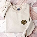 Sand crewneck sweatshirt laid flat on wood, embroidered with a small shaggy golden doodle face on the left chest