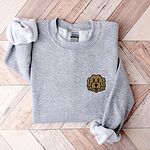 Sport Grey crewneck sweatshirt laid flat on wood, embroidered with a small shaggy golden doodle face on the left chest