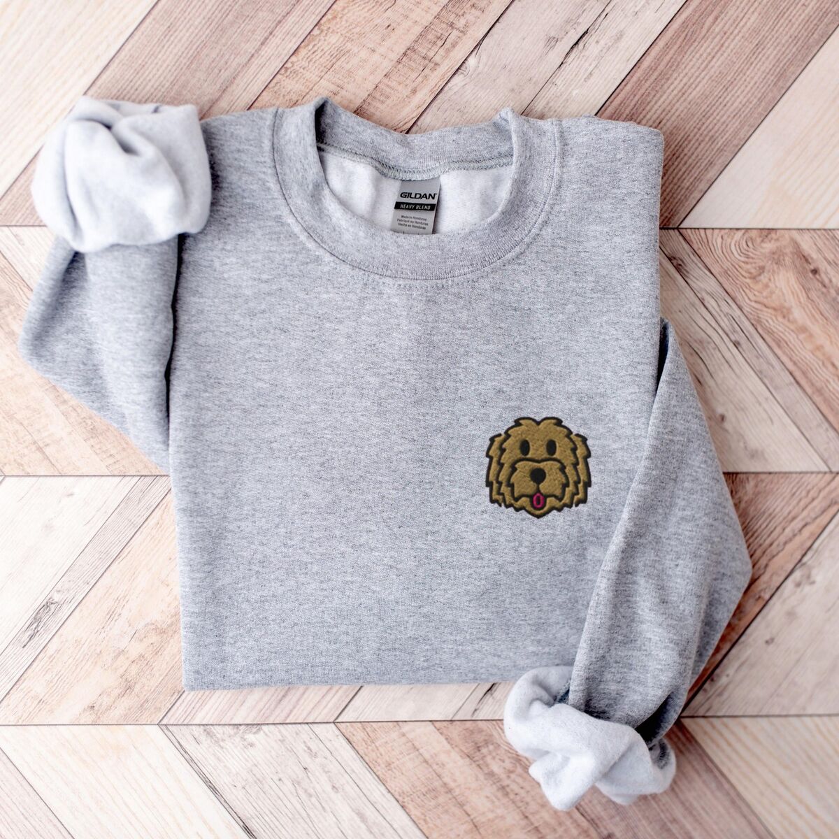 Goldendoodle Face Sweatshirt - Sport Grey Sport Grey crewneck sweatshirt laid flat on wood, embroidered with a small shaggy golden doodle face on the left chest