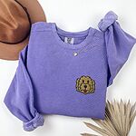 Violet crewneck sweatshirt laid flat with a hat and dried palm leaves, embroidered with a small shaggy golden doodle face on the left chest