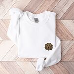 White crewneck sweatshirt laid flat on wood, embroidered with a small shaggy golden doodle face on the left chest
