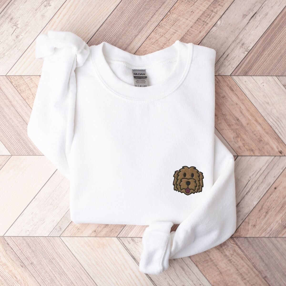 Goldendoodle Face Sweatshirt - White White crewneck sweatshirt laid flat on wood, embroidered with a small shaggy golden doodle face on the left chest