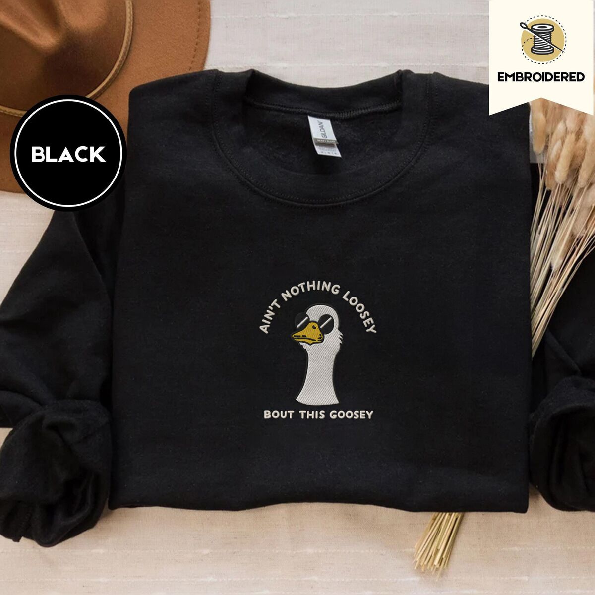 Goose with Sunglasses Sweatshirt – Black Black crewneck sweatshirt with a brown felt hat and dried bunny tail grass, embroidered with a cool white goose in black sunglasses with arched white lettering embroidered on the chest