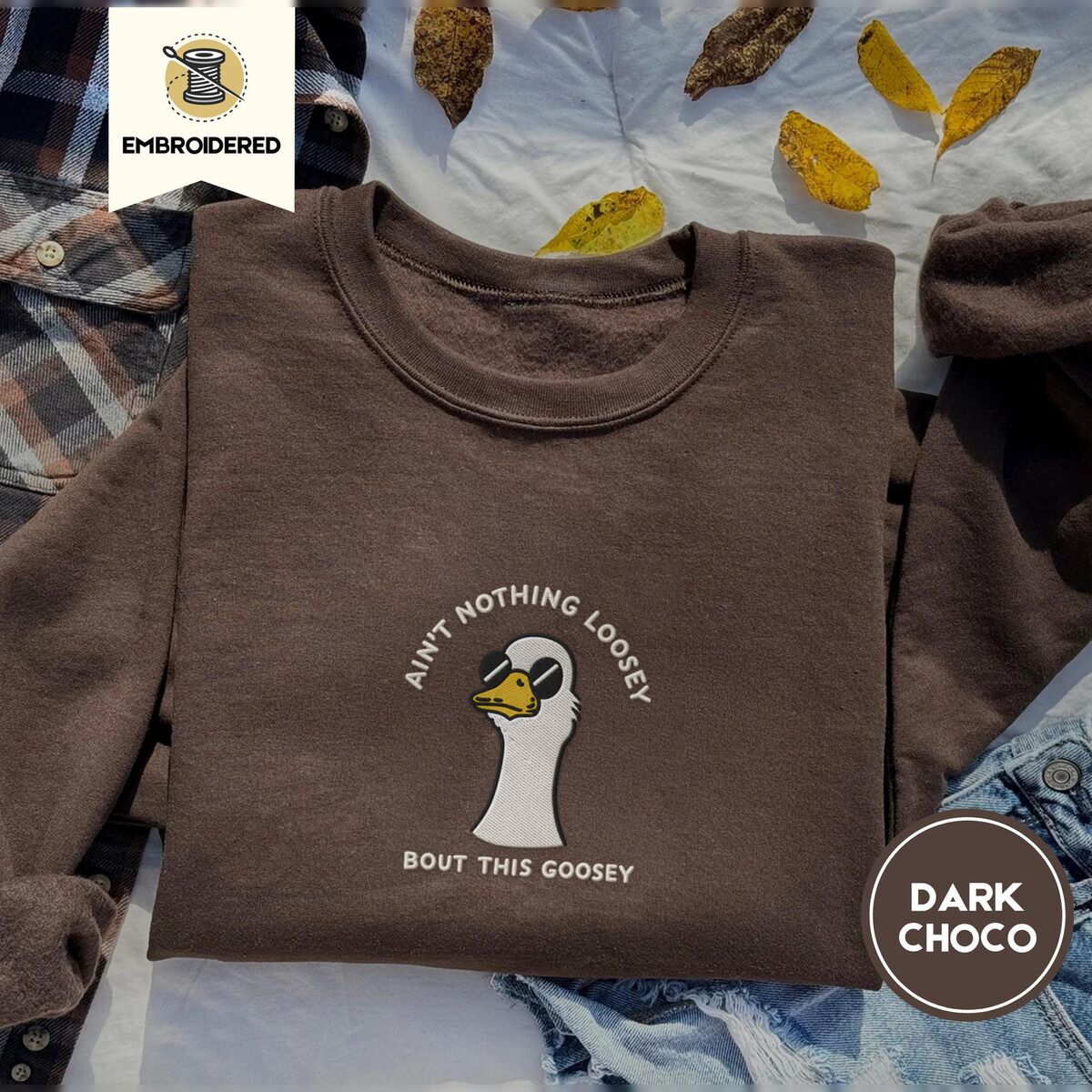Goose with Sunglasses Sweatshirt – Dark Chocolate Dark Chocolate crewneck sweatshirt on a plaid flannel shirt with yellow autumn leaves and denim shorts, embroidered with a cool white goose in black sunglasses with arched white lettering embroidered on the chest