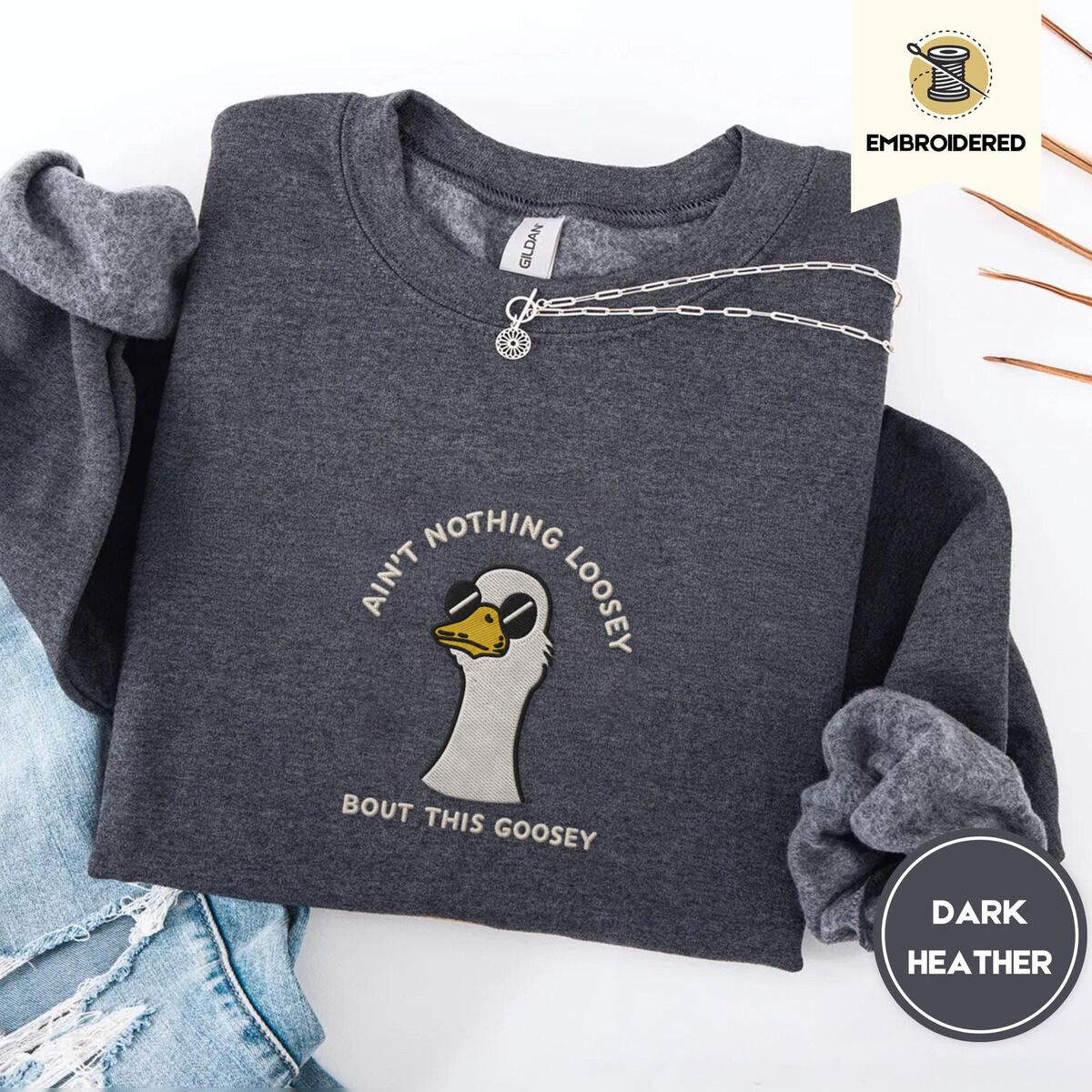 Goose with Sunglasses Sweatshirt – Dark Heather Dark Heather crewneck sweatshirt folded with ripped jeans and a silver chain necklace, embroidered with a cool white goose in black sunglasses with arched white lettering embroidered on the chest