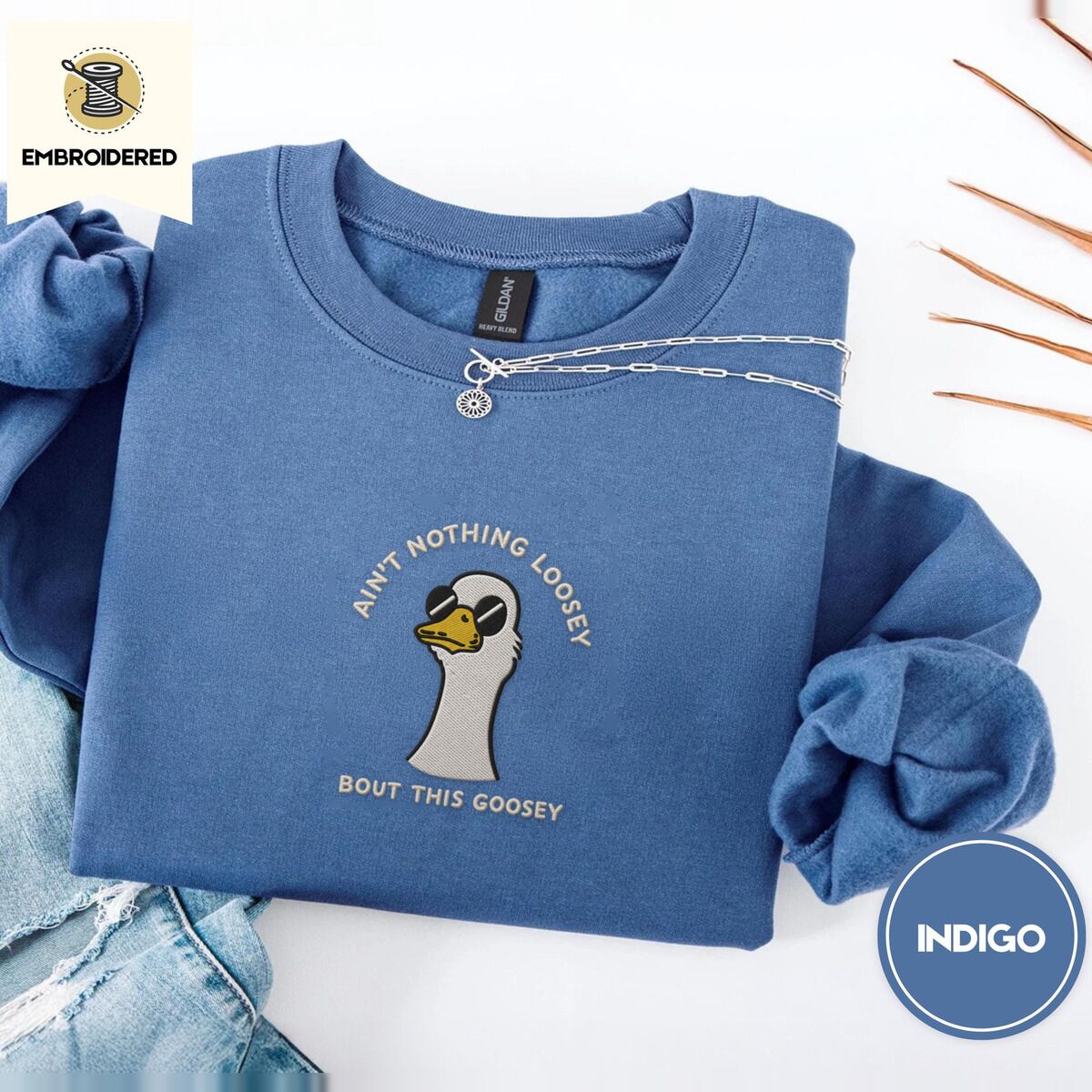 Goose with Sunglasses Sweatshirt – Indigo Blue Indigo Blue crewneck sweatshirt folded with light jeans and dried twigs, embroidered with a cool white goose in black sunglasses with arched white lettering embroidered on the chest