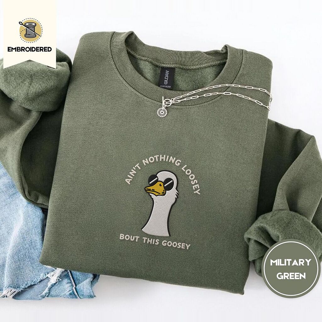 Military Green crewneck sweatshirt folded with light jeans and a silver chain necklace, embroidered with a cool white goose in black sunglasses with arched white lettering embroidered on the chest