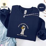 Navy crewneck sweatshirt folded with ripped jeans and a silver chain necklace, embroidered with a cool white goose in black sunglasses with arched white lettering embroidered on the chest