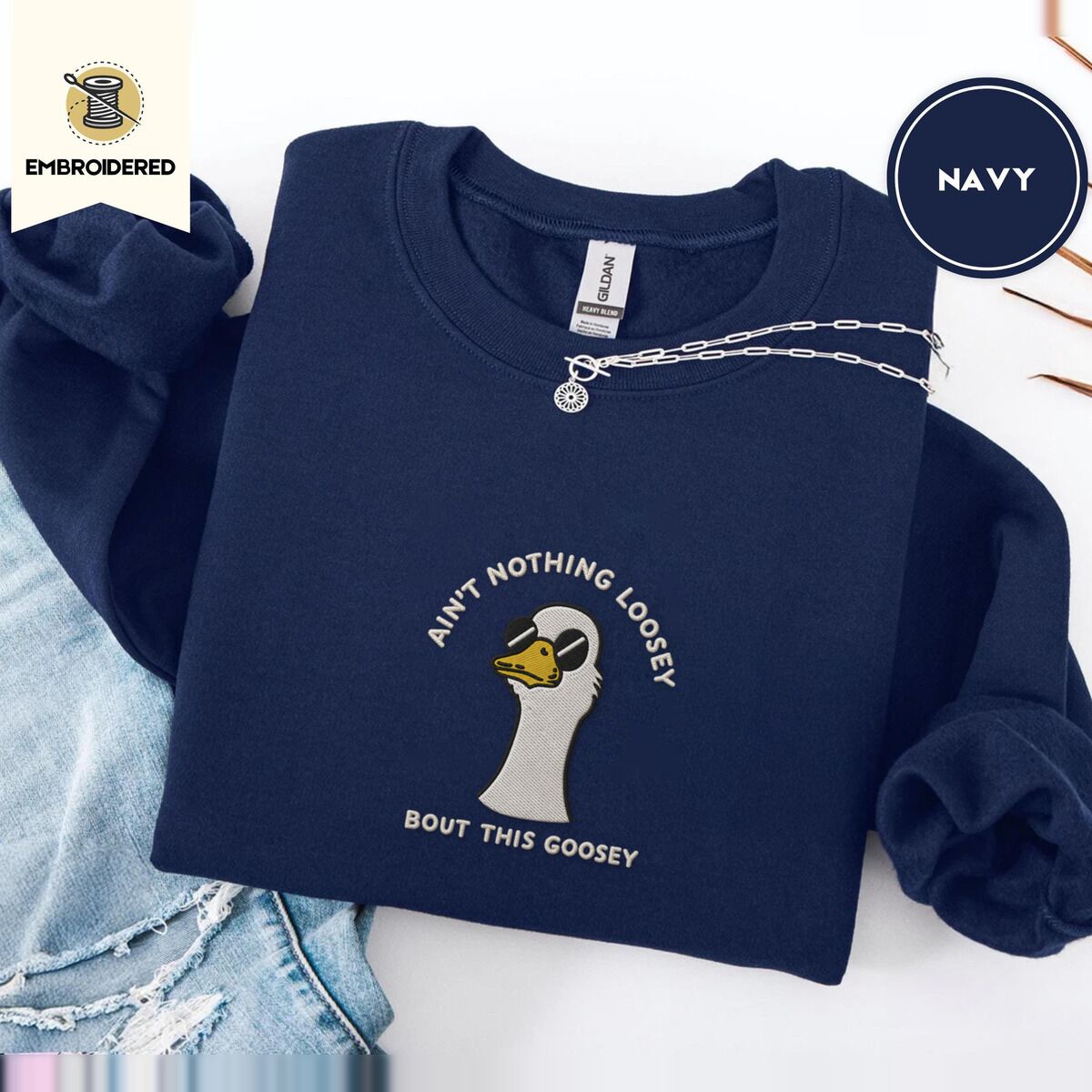 Goose with Sunglasses Sweatshirt – Navy Navy crewneck sweatshirt folded with ripped jeans and a silver chain necklace, embroidered with a cool white goose in black sunglasses with arched white lettering embroidered on the chest