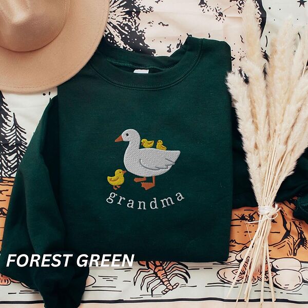 Forest Green crewneck sweatshirt folded with a tan felt hat and pampas grass, embroidered with a white mama duck, yellow ducklings and the word grandma in white thread
