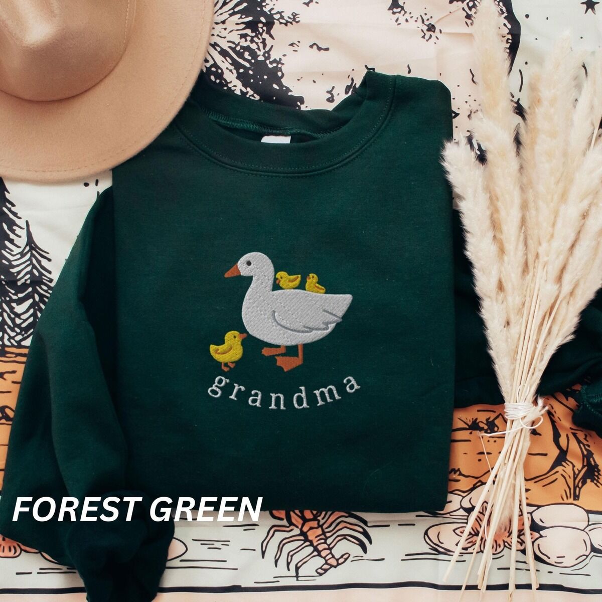 Grandma Duck Sweatshirt – Forest Green Forest Green crewneck sweatshirt folded with a tan felt hat and pampas grass, embroidered with a white mama duck, yellow ducklings and the word grandma in white thread