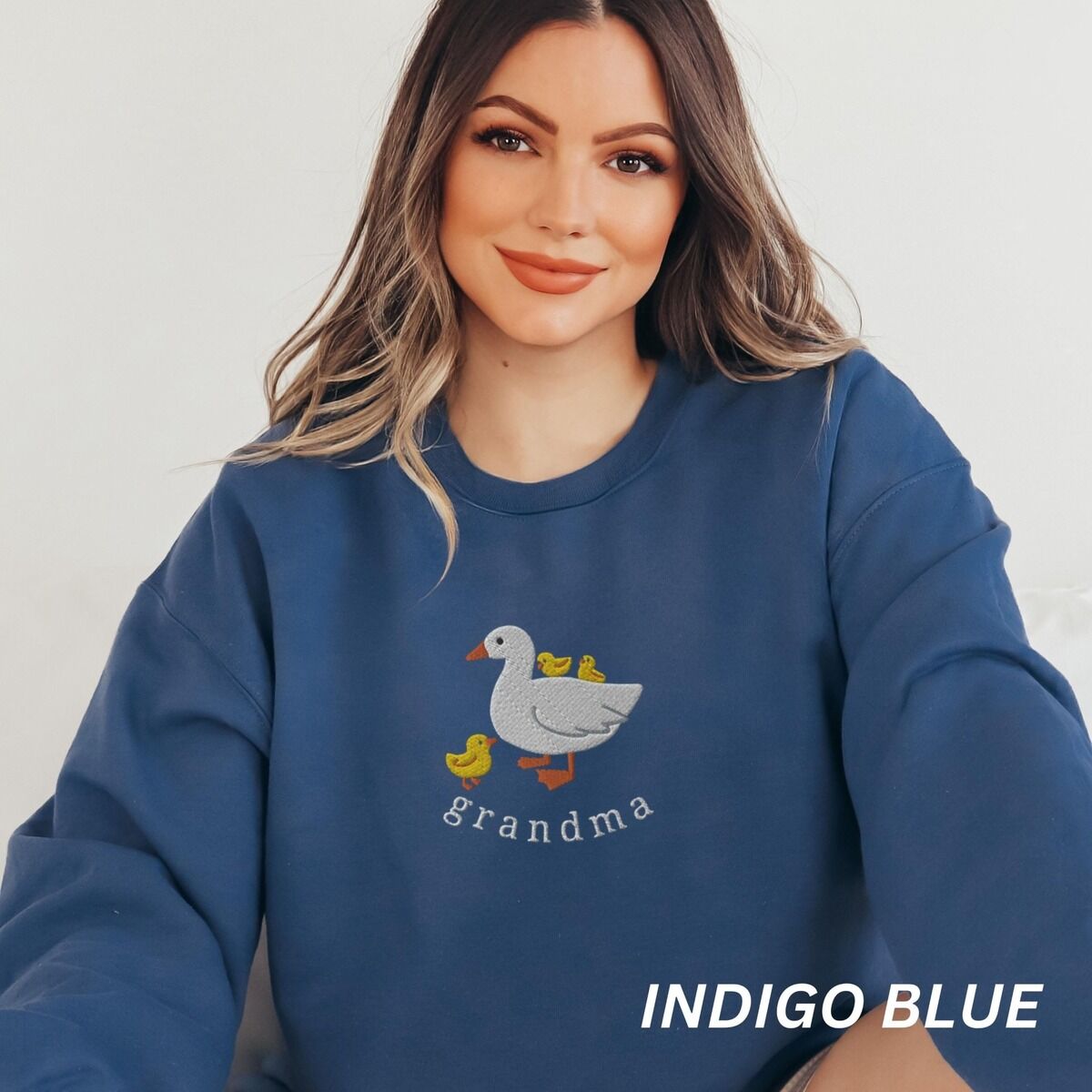Grandma Duck Sweatshirt – Indigo Blue Woman wearing an Indigo Blue crewneck sweatshirt embroidered with a white mama duck, yellow ducklings and the word grandma in white thread