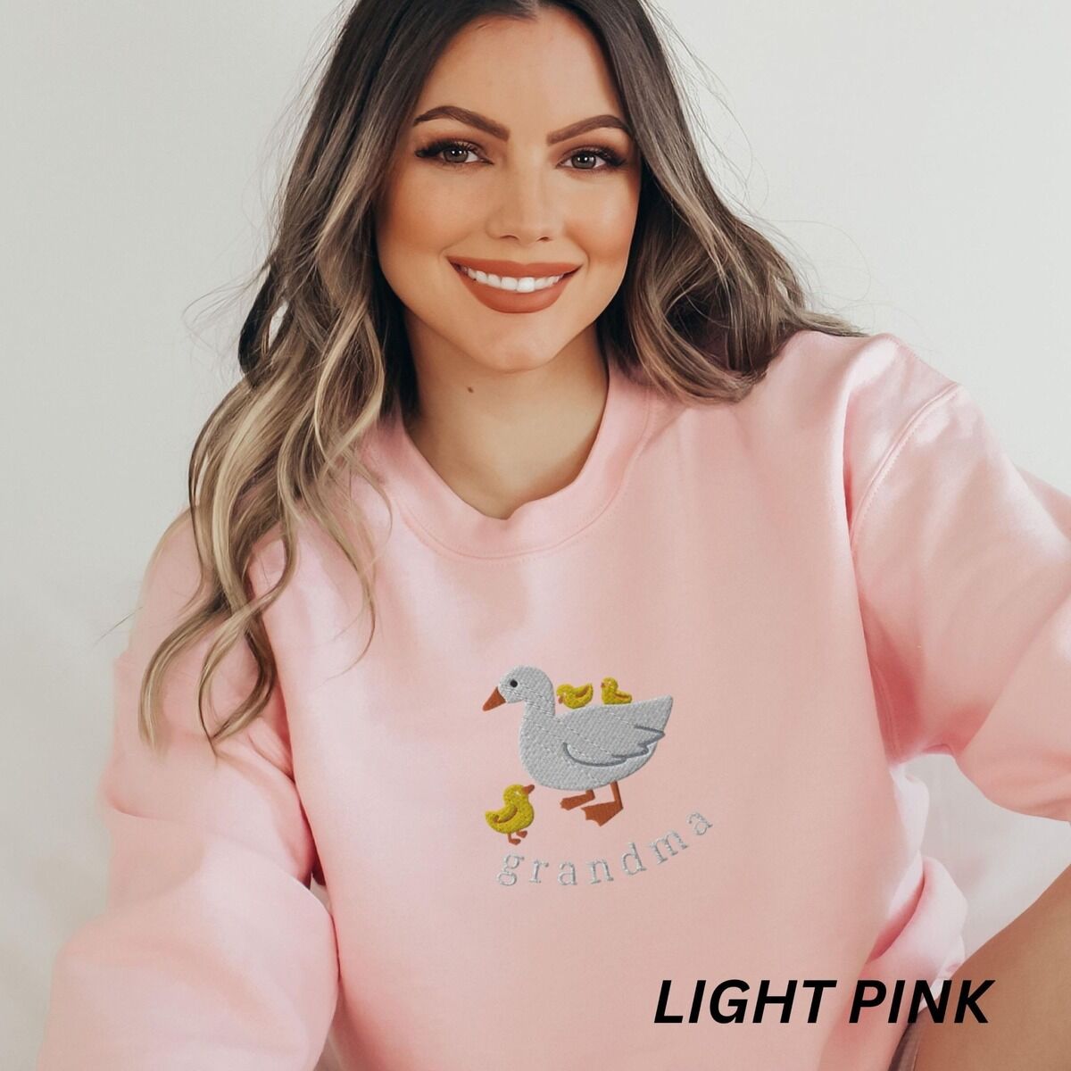 Grandma Duck Sweatshirt – Light Pink Woman wearing a Light Pink crewneck sweatshirt embroidered with a white mama duck, yellow ducklings and the word grandma in white thread