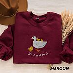 Maroon crewneck sweatshirt folded with a brown felt hat and dried grass, embroidered with a white mama duck, yellow ducklings and the word grandma in white thread