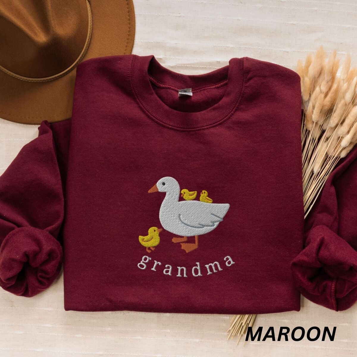 Grandma Duck Sweatshirt – Maroon Maroon crewneck sweatshirt folded with a brown felt hat and dried grass, embroidered with a white mama duck, yellow ducklings and the word grandma in white thread