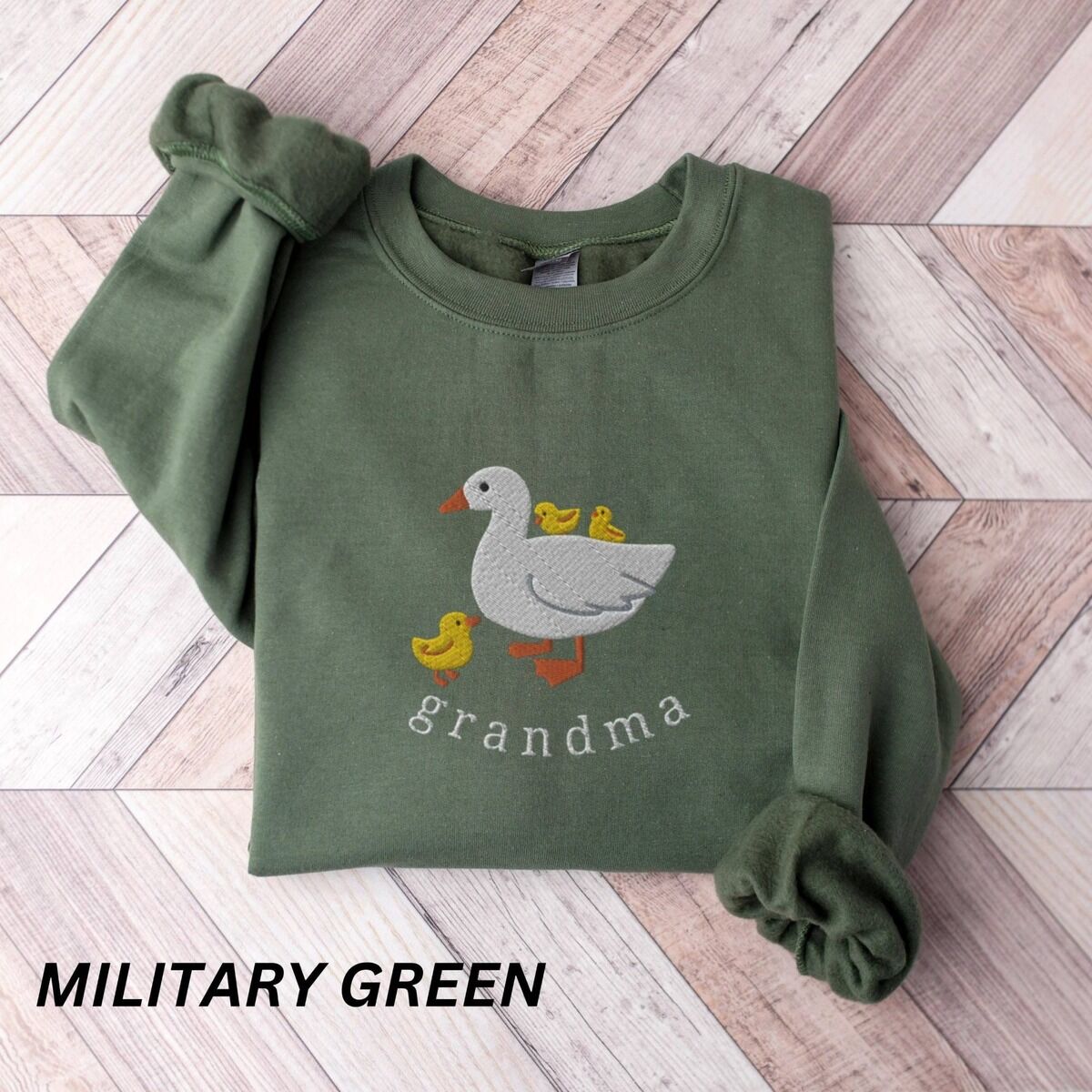 Grandma Duck Sweatshirt – Military Green Military Green crewneck sweatshirt folded on a wooden floor, embroidered with a white mama duck, yellow ducklings and the word grandma in white thread