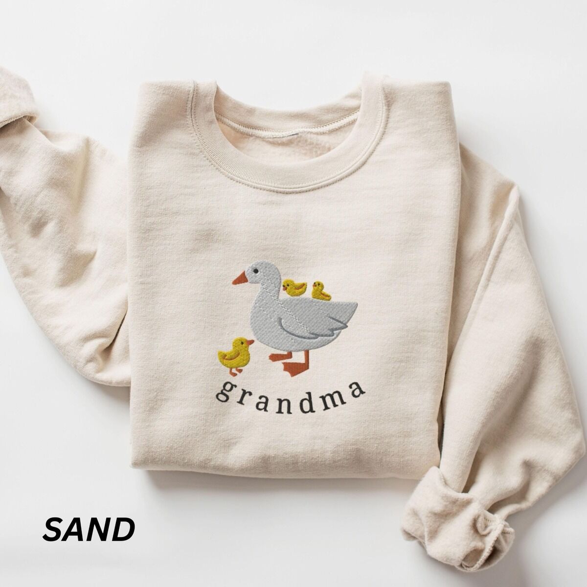 Grandma Duck Sweatshirt – Sand Sand crewneck sweatshirt folded on a white background, embroidered with a white mama duck, yellow ducklings and the word grandma in black thread