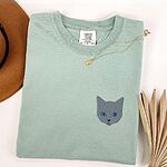 Bay T-shirt laid flat beside a tan hat with a gold necklace, with a gray cat face with blue eyes embroidered on the left chest
