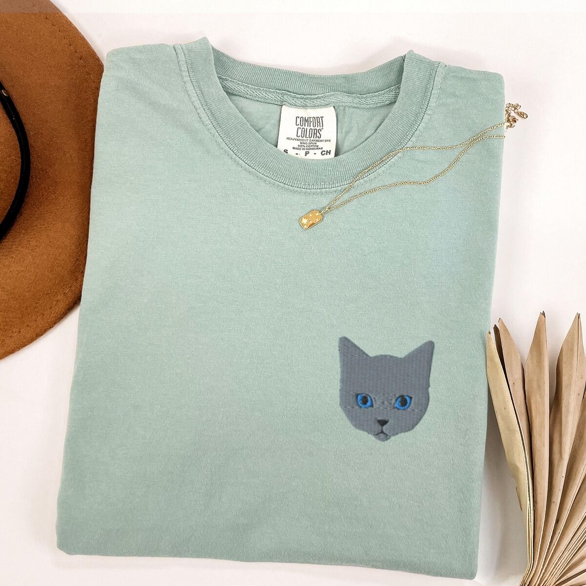 Grey Cat Shirt – Bay Bay T-shirt laid flat beside a tan hat with a gold necklace, with a gray cat face with blue eyes embroidered on the left chest