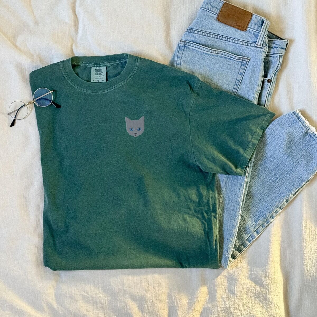 Grey Cat Shirt – Blue Spruce Blue Spruce T-shirt laid on a bed beside jeans and glasses, with a gray cat face with blue eyes embroidered on the left chest