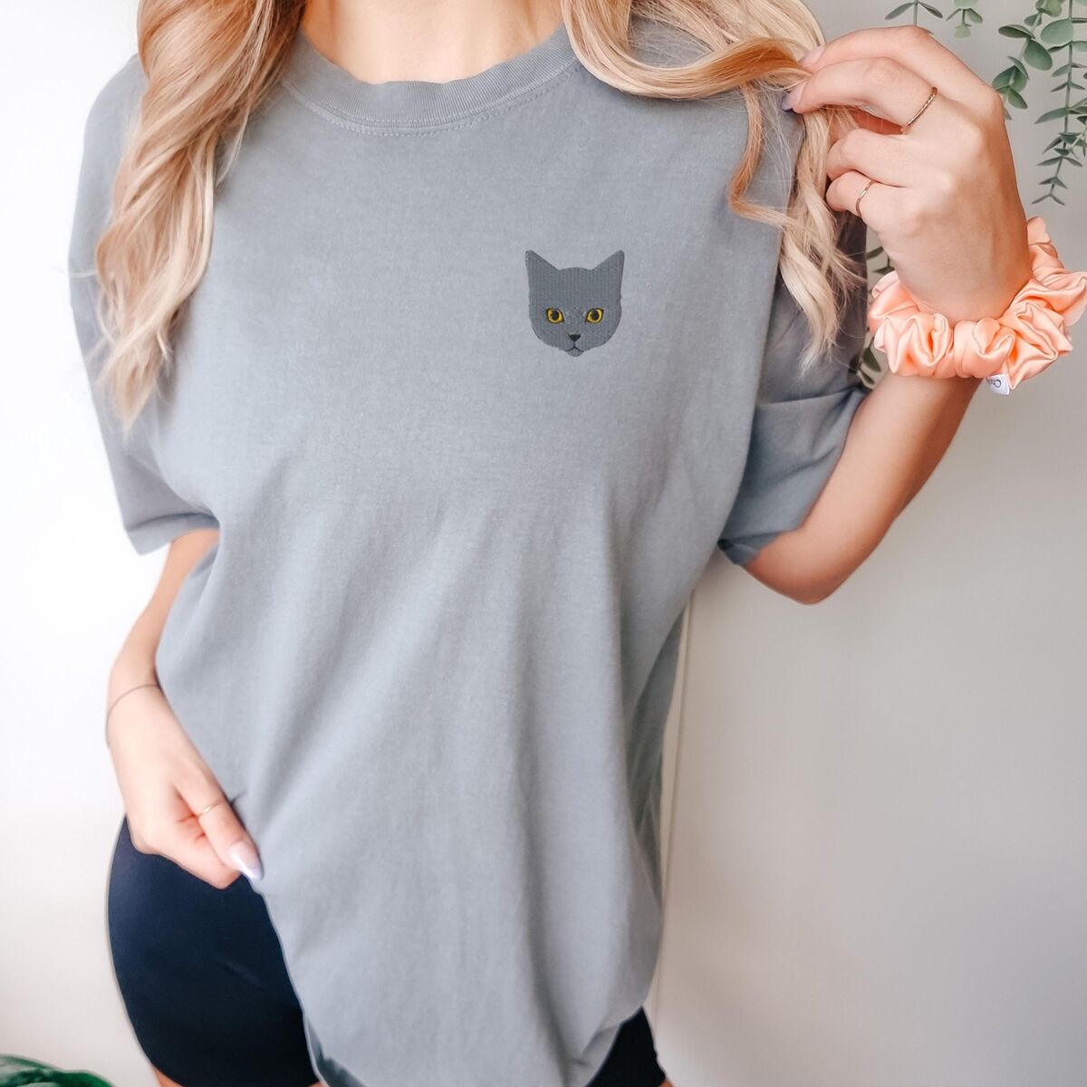Grey Cat Shirt Grey Woman with an orange scrunchie in a Grey T-shirt with a gray cat face with gold eyes embroidered on the left chest