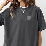 Woman with a gold necklace in a Pepper T-shirt with a gray cat face with gold eyes embroidered on the left chest