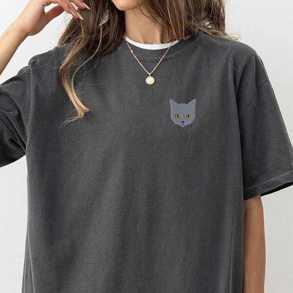 Woman with a gold necklace in a Pepper T-shirt with a gray cat face with gold eyes embroidered on the left chest