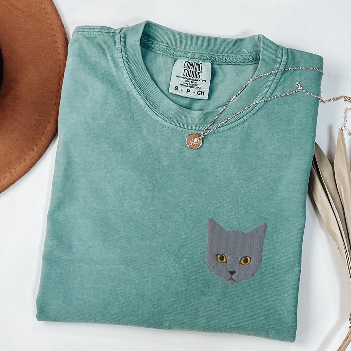 Grey Cat Shirt – Seafoam Seafoam T-shirt laid flat with a gold necklace, with a gray cat face with gold eyes embroidered on the left chest