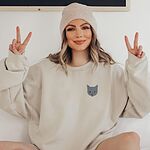 Smiling woman in a knit beanie and a Sand crewneck sweatshirt making peace signs, with a gray cat face with gold eyes embroidered on the left chest