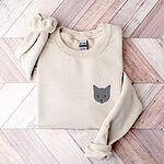 Sand grey cat sweatshirt folded on a pink and white herringbone wood floor, with a gray cat face with gold eyes embroidered on the left chest