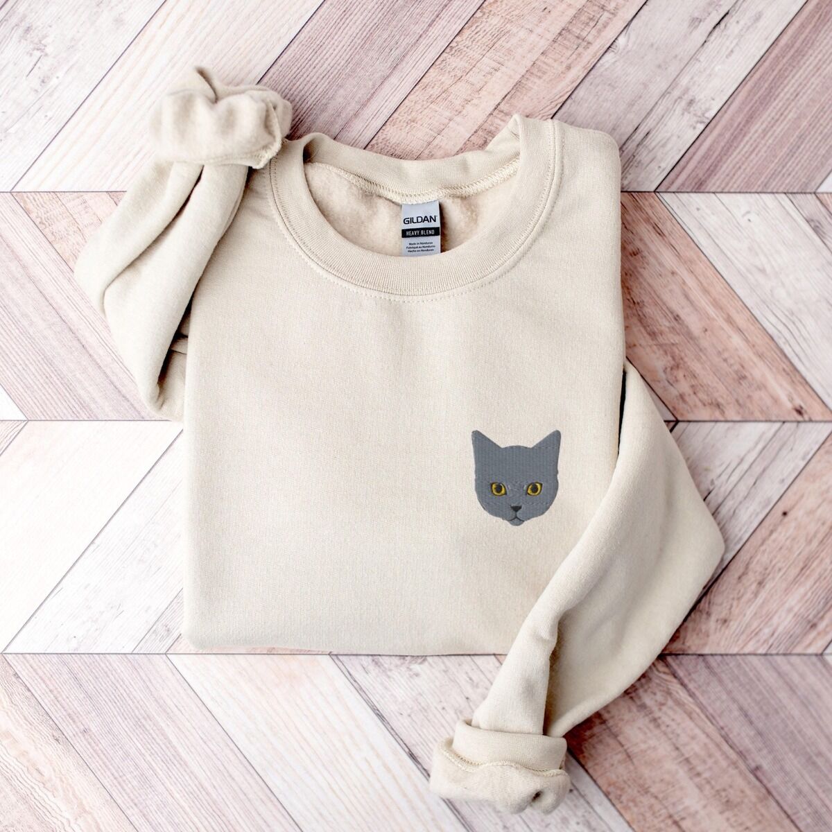 Grey Cat Sweatshirt – Sand Sand grey cat sweatshirt folded on a pink and white herringbone wood floor, with a gray cat face with gold eyes embroidered on the left chest