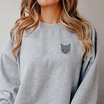 Woman with long wavy hair in a Sport Grey crewneck sweatshirt with a gray cat face with gold eyes embroidered on the left chest